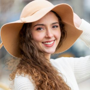 Smiling woman with curly hair and a stylish hat outdoors, exuding warmth and happiness.